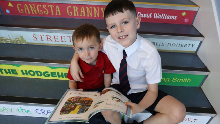 Two young boys, one a nursery pupil, sit together on a set of stairs bearing names of famous children's authors as they read a book together.