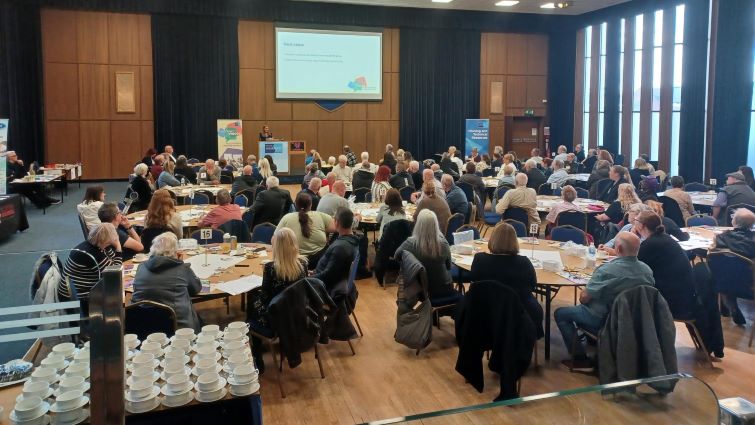 This is a general view of the Customer Voices Conference held in the council's banqueting hall, with people sat around tables as a presentation is made on the stage