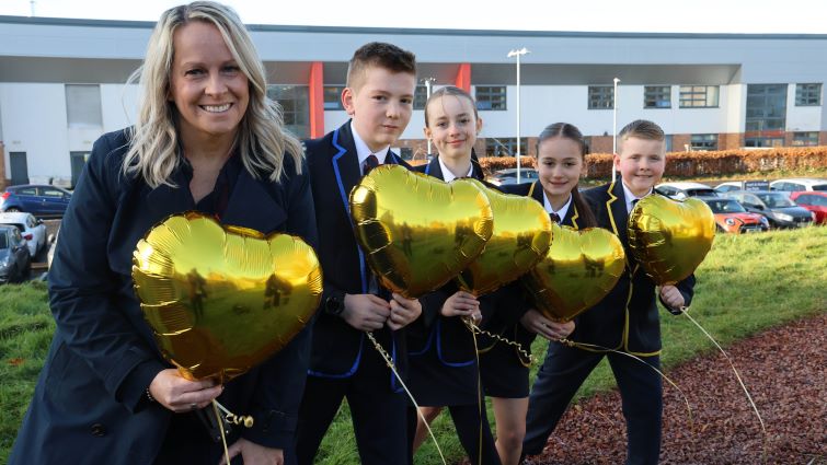 Head Teacher Isabelle Murray and four pupils are pictured in a line standing outside the school holiding five gold-coloured, heart-shaped balloons.