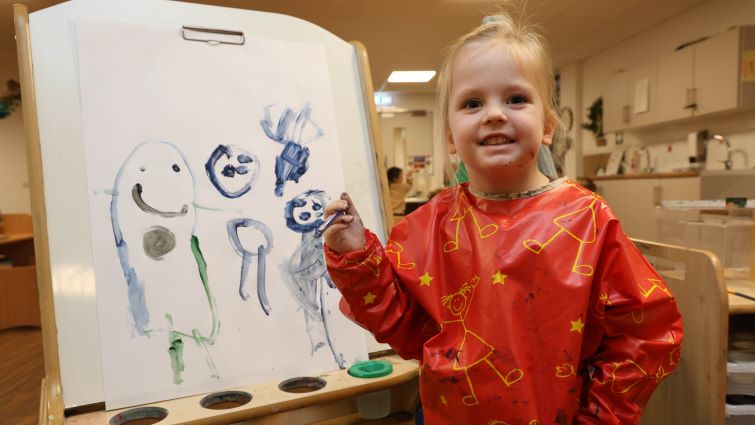 This image shows a nursery pupil holding a paint brush standing in front of a picture she has painted
